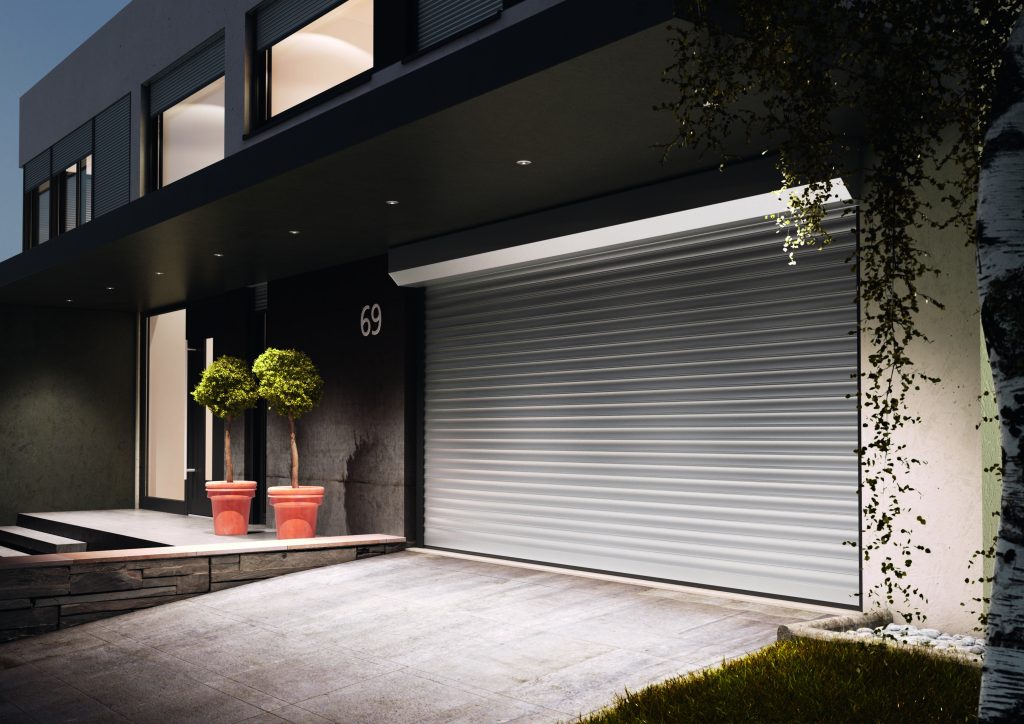 Modern house at night with number 69, white roller garage door, two potted topiary trees, and stone driveway entrance