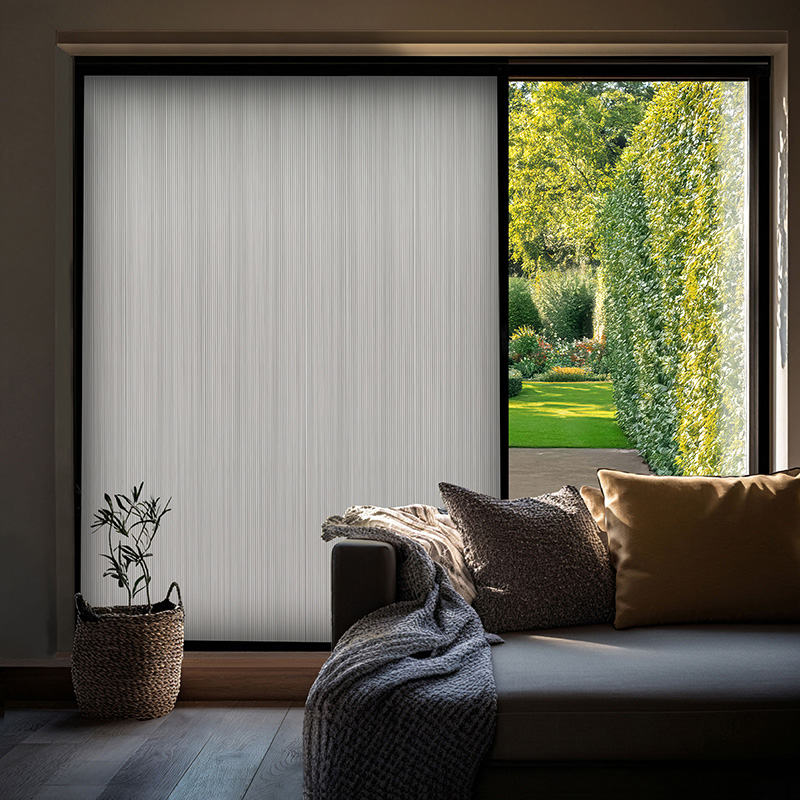 Sliding glass door with textured cellular shade half-closed, sofa with pillows, view of garden with hedges and flowers.