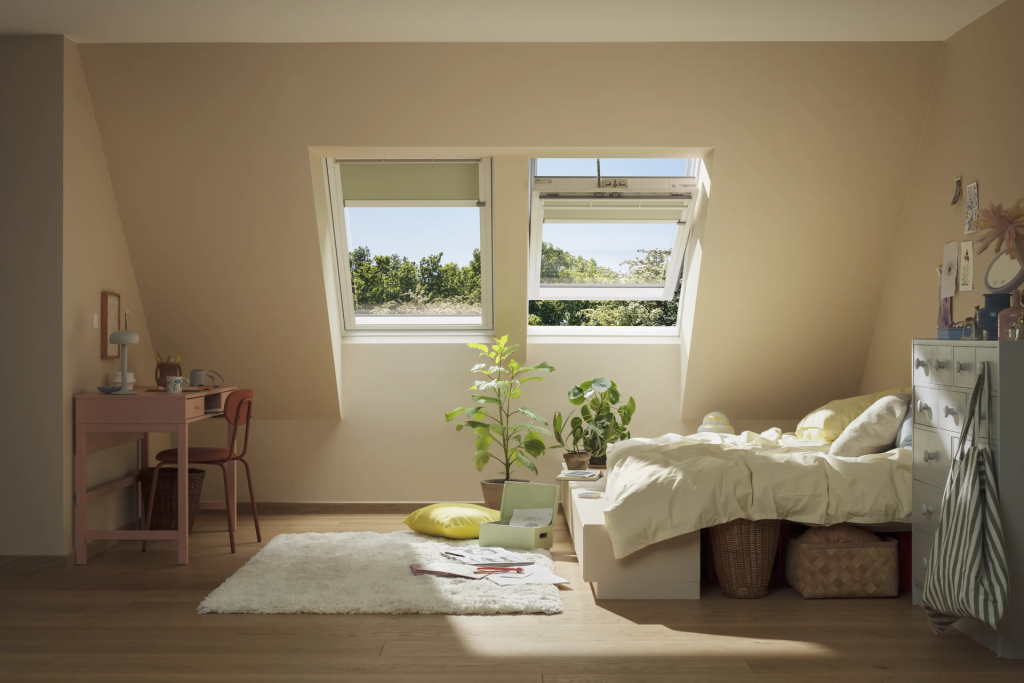 Cozy attic bedroom with skylight windows, desk, plants, white rug, and unmade bed with storage baskets underneath.