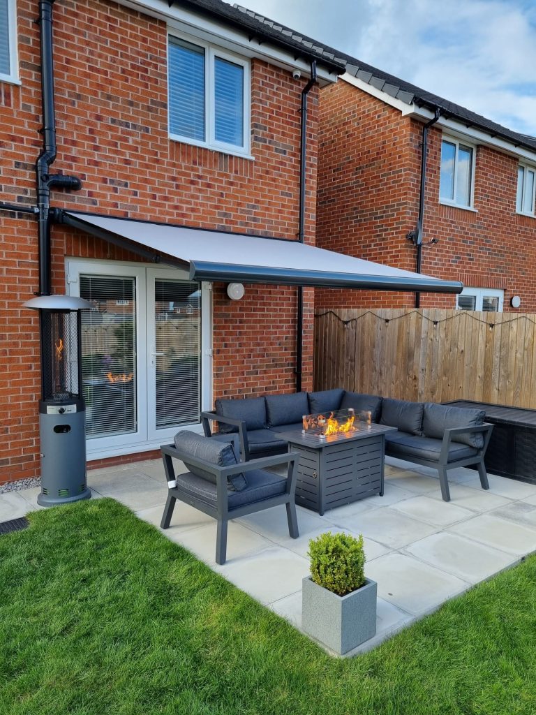 Grey outdoor patio set with fire pit table under retractable awning on brick house patio with lawn