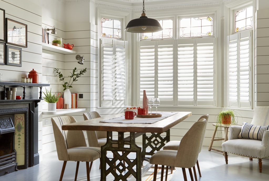 Bay window dining room with white plantation shutters, stained glass, wood table, and vintage cast iron fireplace.