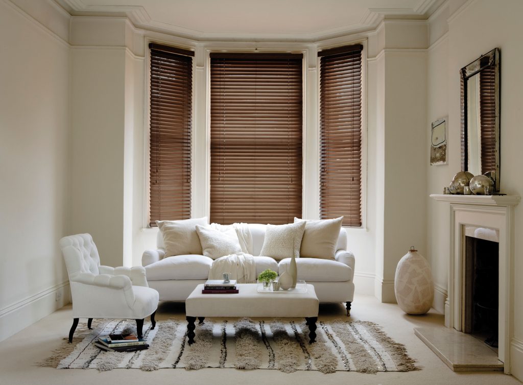 Cream living room with bay window wooden blinds, white sofa, armchair, tufted ottoman, and fireplace mantel decor.