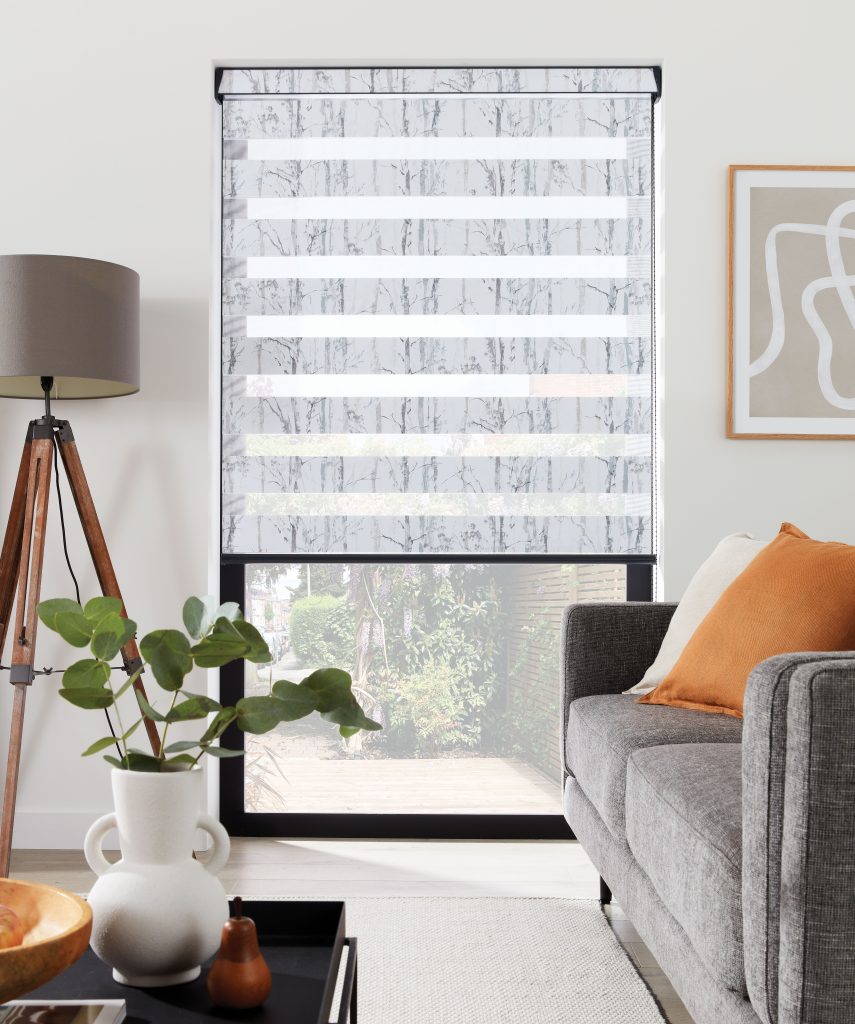 Living room with tree-patterned day-night blind on window, gray sofa with orange pillow, tripod lamp, and plant decor.