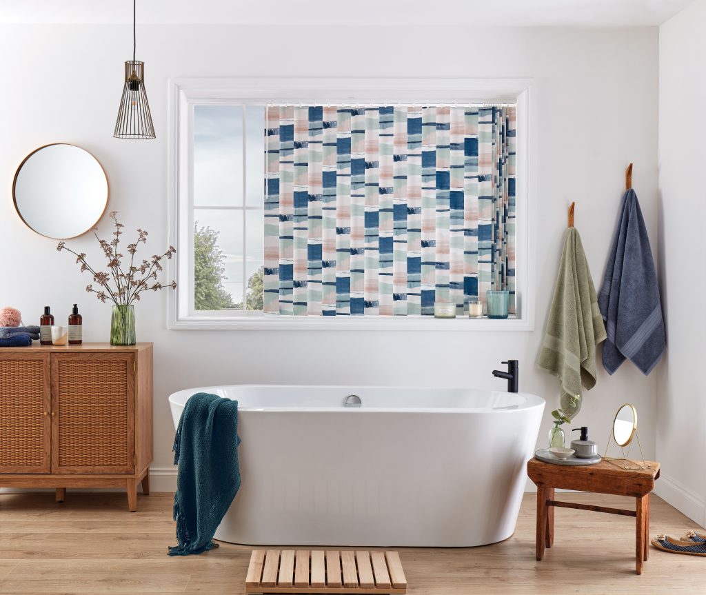 Freestanding bathtub with patterned vertical blinds, wooden vanity, hanging towels, and round mirror in modern bathroom decor.