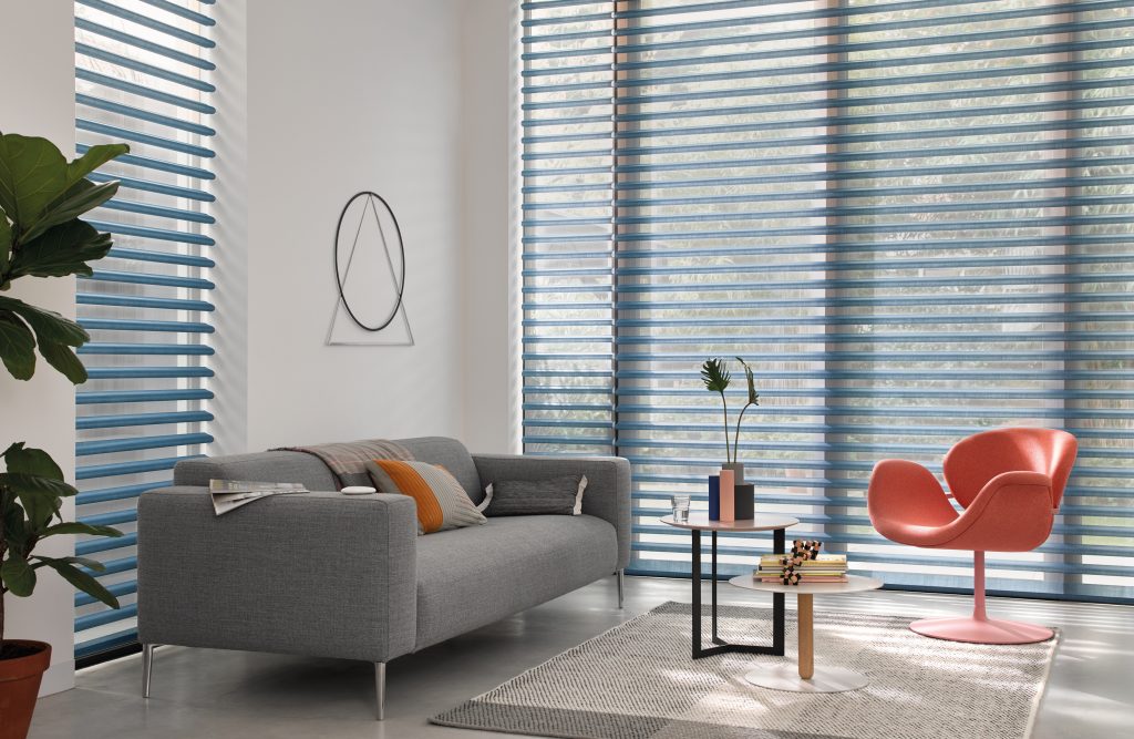 Modern living room with blue sheer window blinds, gray sofa, coral chair, and round side tables on a woven rug.