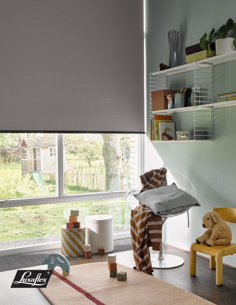 Grey Luxaflex roller blind in children's playroom with shelving, teddy bear, and garden view through window.