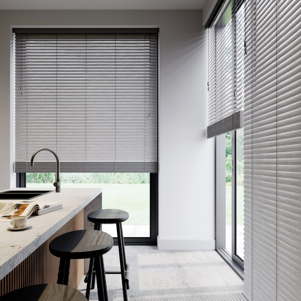 Modern kitchen with concrete island, black bar stools, and gray venetian blinds on large windows overlooking green garden.
