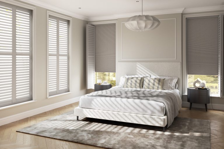Elegant bedroom with upholstered bed, striped pillows, plantation shutters, gray cellular shades, and light wood floors.