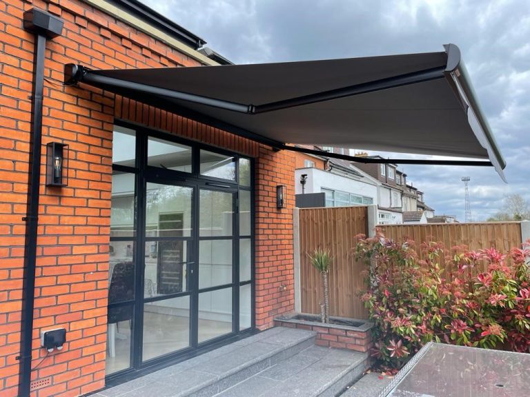 Retractable dark grey patio awning on red brick house with black-framed glass doors, overlooking garden with red-leaved plants.