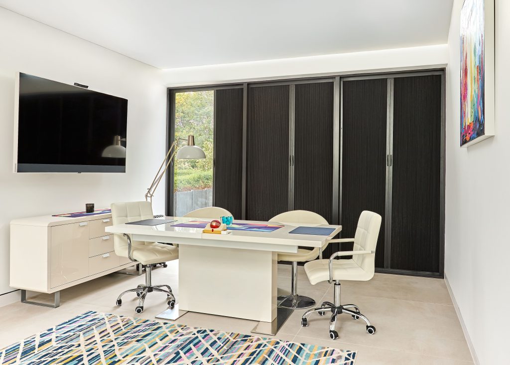 Modern home office with white desk, cream leather chairs, wall-mounted TV, colorful geometric rug, and abstract art