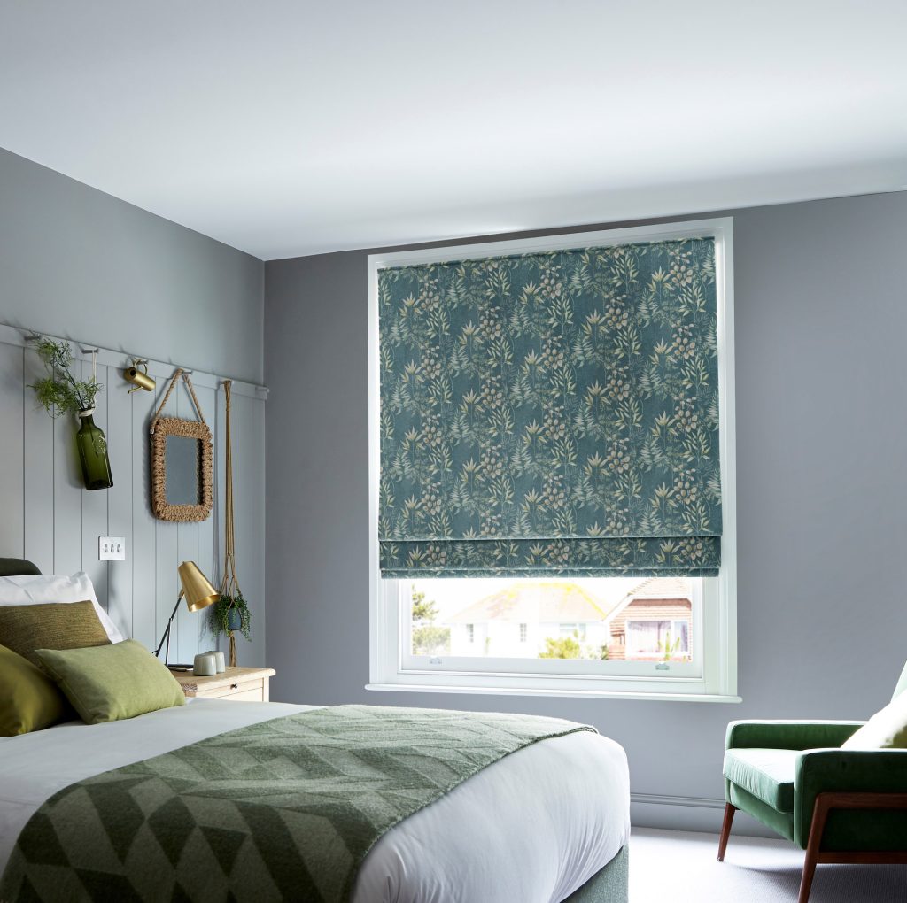 Bedroom with teal floral Roman blind, grey walls, green cushions, wicker mirror, and gold accent decor.