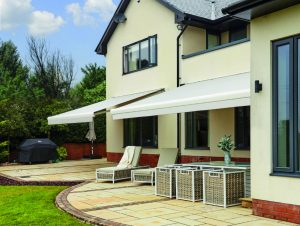 Cream retractable patio awning over rattan furniture on stone patio with landscaped garden and BBQ grill cover