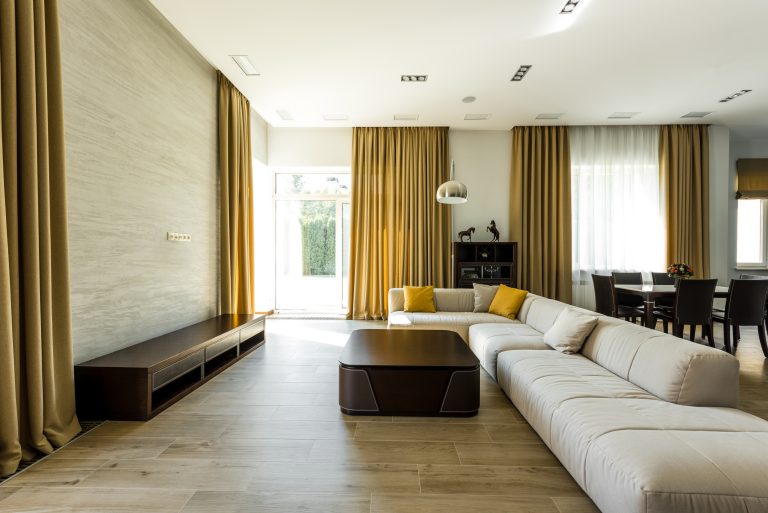 Modern living room with beige sectional sofa, mustard curtains, dark wood coffee table, and dining area with wood floors.