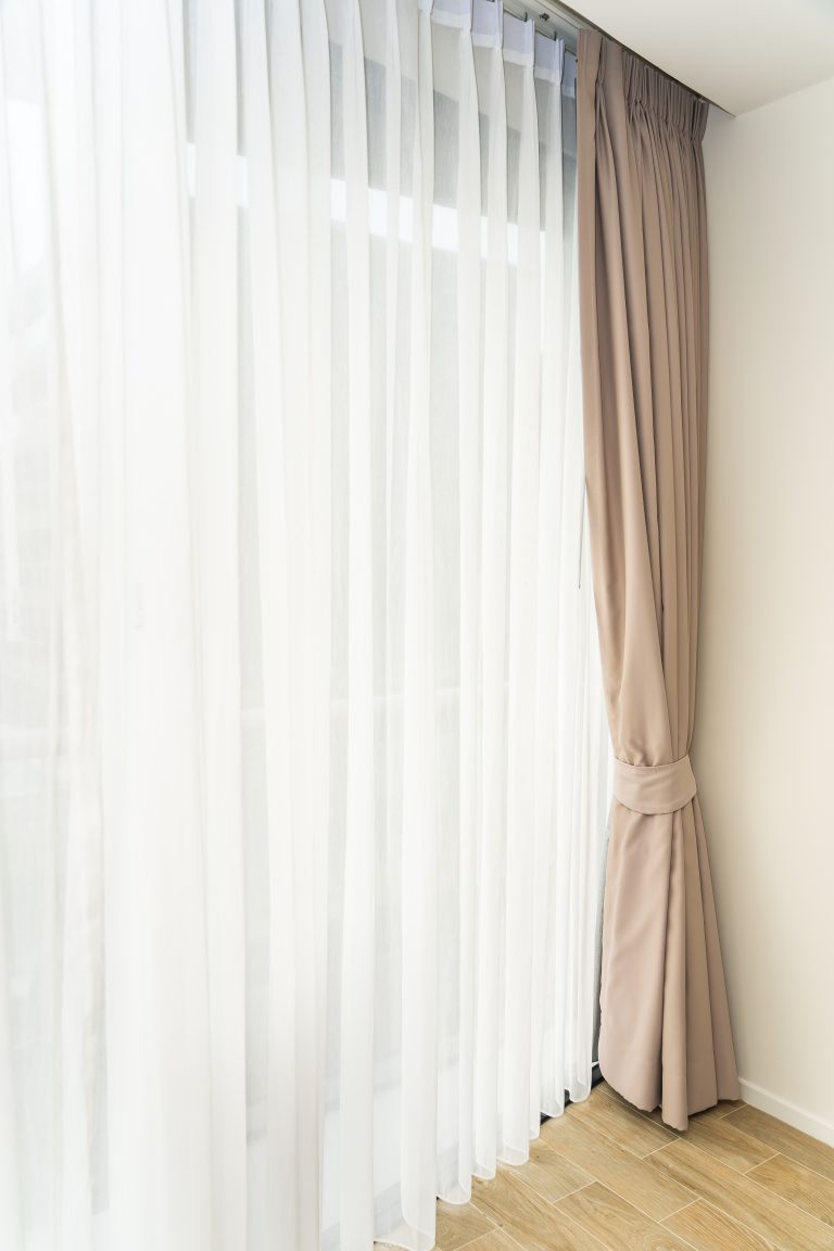 Sheer white curtains and beige drapery tied back beside a large window with wooden flooring